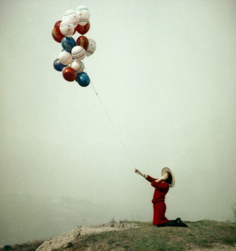 Giovane donna in abito rosso e cappello, inginocchiata, tiene il filo con alcuni palloncini colorati.1970 - 1980